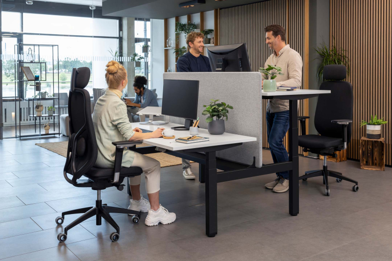Drei Kollegen im Büro stehen und sitzen an höhenverstellbaren Schreibtischen