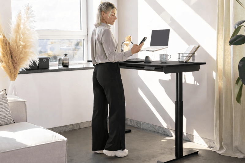 Eine blonde Frau steht an einem schwarzen, höhenverstellbaren Schreibtisch in einem hellen Home-Office und schaut auf ihr Smartphone. Auf der Tischplatte steht ein Laptop auf einem Ständer