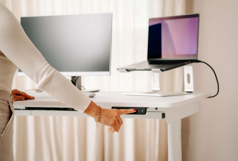 Nahaufnahme der Hand einer Frau, die das elektrische Bedienfeld eines weißen höhenverstellbaren Schreibtischs bedient. Auf der Tischplatte stehen Monitor und Laptop
