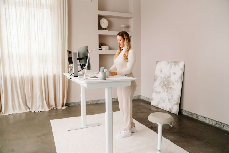 Eine Frau arbeitet stehend an einem modernen, höhenverstellbaren Schreibtisch weiß mit Dual-Monitor-Setup in einem hellen, minimalistischen Home-Office. Rechts ist ein weißer ergonomischer Wipphocker zu sehen